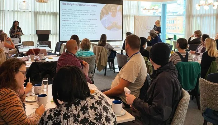 People in a meeting room, sat at tables looking towards a projector screen