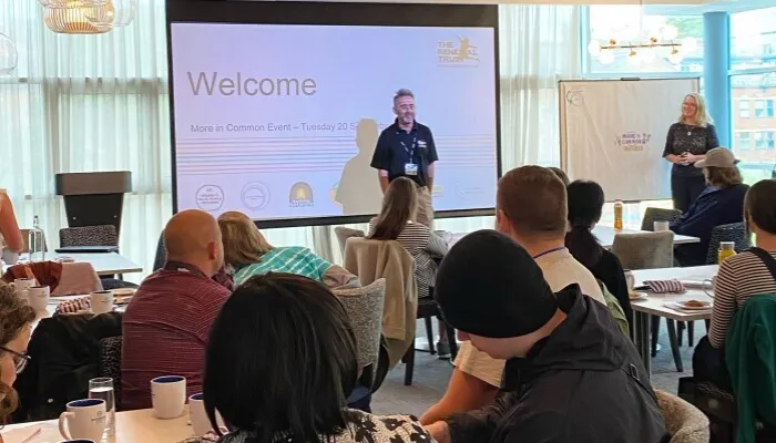 People in a meeting room, sat at tables looking towards a person in front of a projector screen