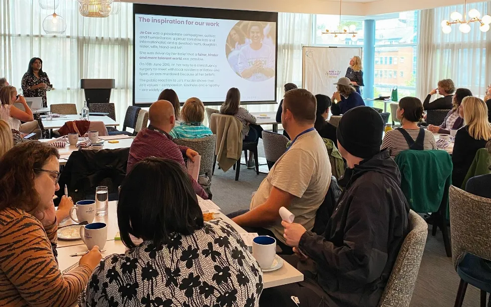 People in a meeting room, sat at tables looking towards a projector screen
